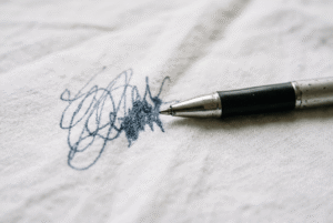 Kugelschreiber-Fleck auf weißem Stoff – ein aufgeklappter Kugelschreiber liegt neben einem frischen Tintenfleck auf hellem Textil.