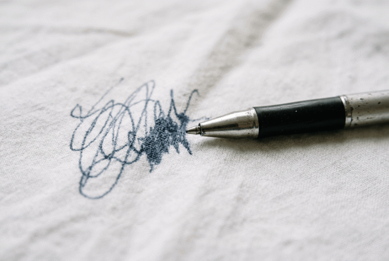 Kugelschreiber-Fleck auf weißem Stoff – ein aufgeklappter Kugelschreiber liegt neben einem frischen Tintenfleck auf hellem Textil.